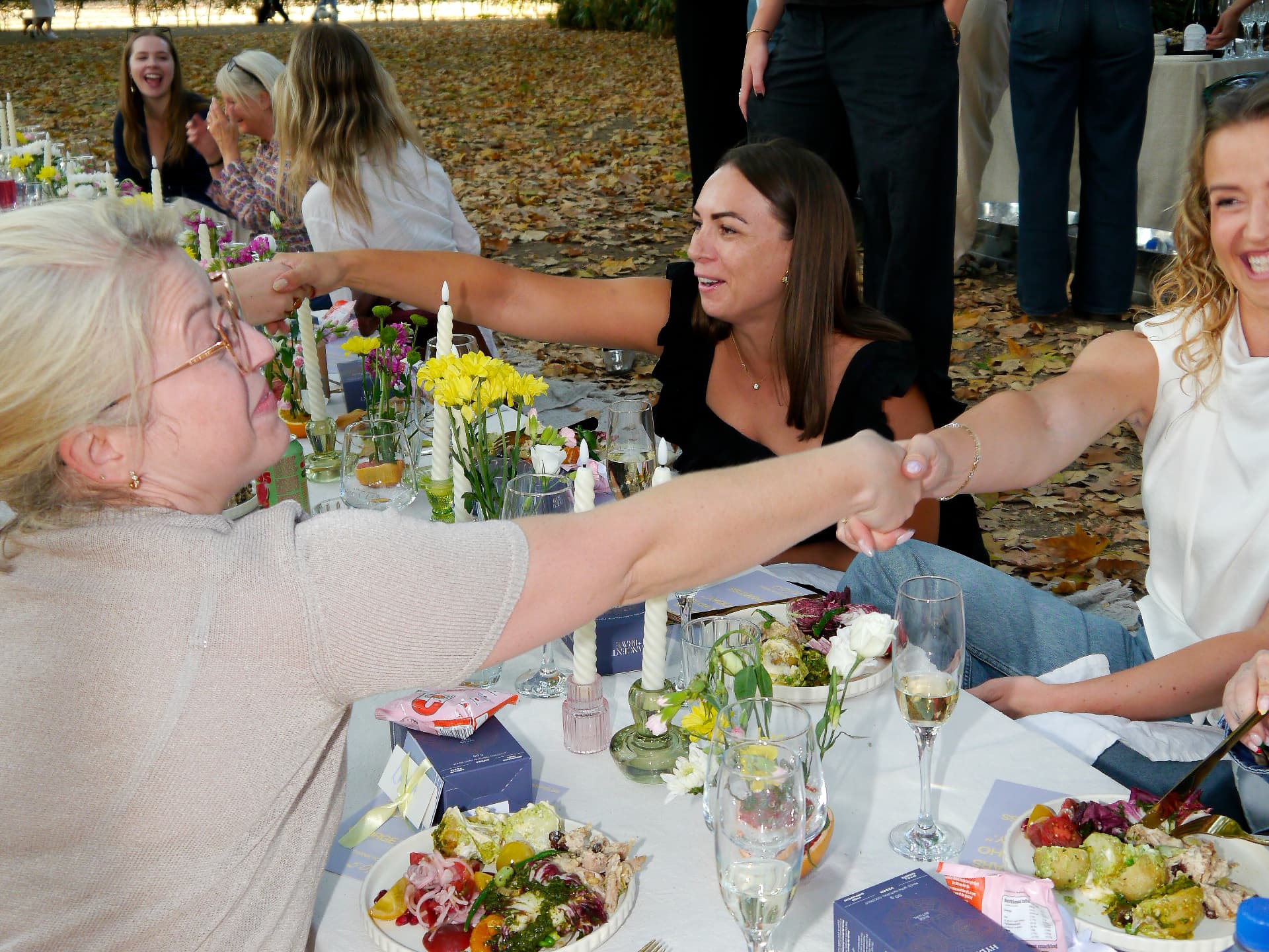 Women seated around an outdoor her.arca table, reaching across to greet one another